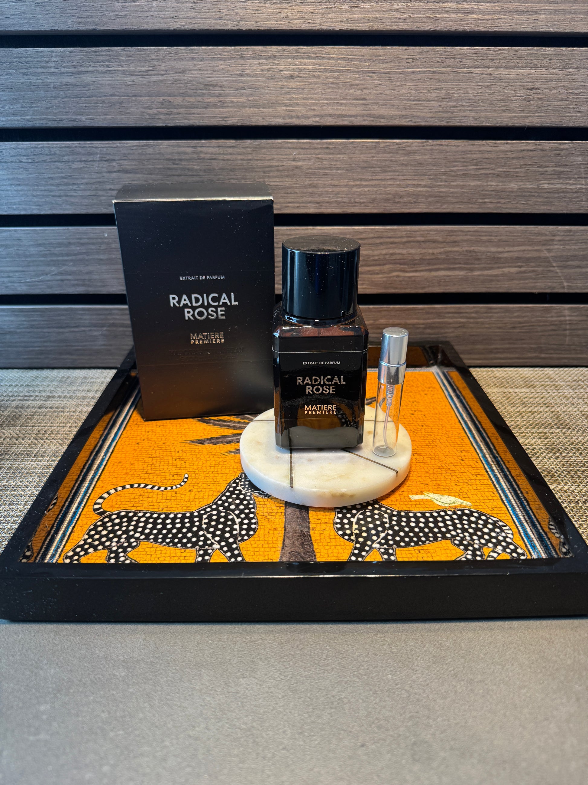 A bottle of matiere Premiere Radical Rose Eau de Parfum with its black packaging box, displayed on a decorative tray with a small travel spray vial against a dark wood slat background.